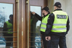 Two police officers knocking on a glass door, before entering without a warrant, due to exigent circumstances.