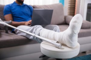 An injured remote worker on his laptop while resting his injured foot on a stool.