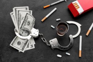 A set of handcuffs strewn on a table next to a shot of whiskey, money, and cigarettes-- representing the potential costs of a DUI.