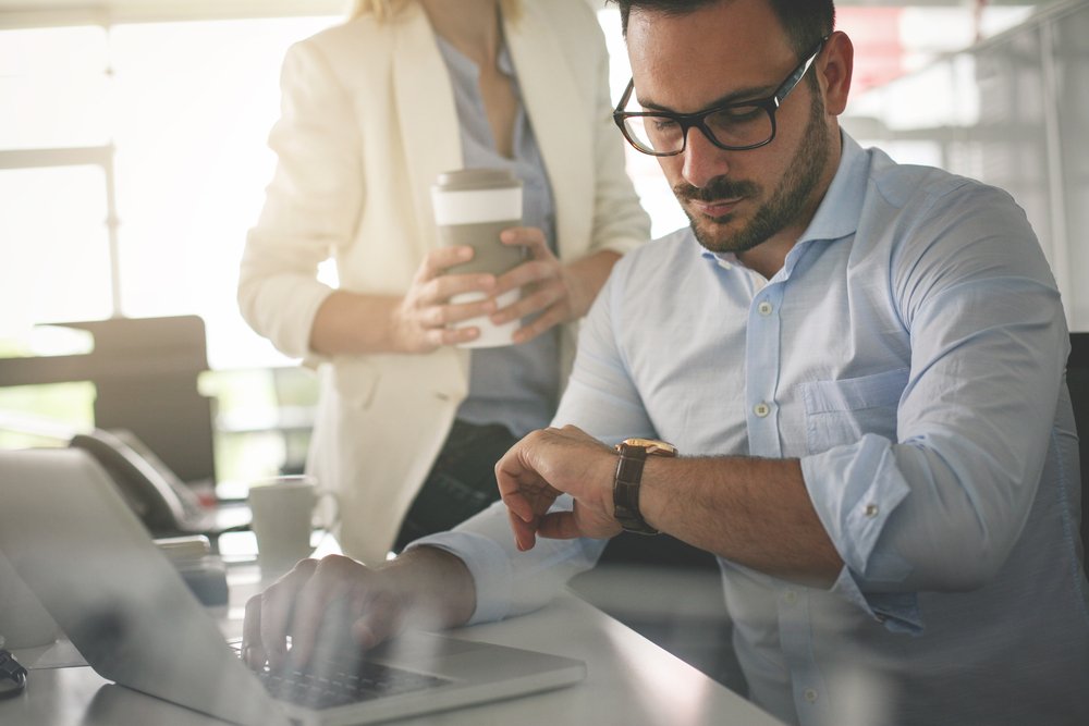 An office worker at his desk and typing on his laptop with one hand while looking at his watch on the other to see how many hours are left in the work day, and there is a co-worker holding coffee behind him