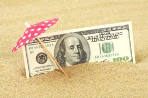 A one hundred dollar bill settled into the sand, under a cocktail umbrella-- representing a paid vacation.