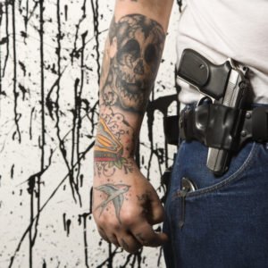 Closeup of man's belt holster carrying a firearm next to his tattooed arm. 