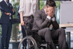 An office worker wearing a suit and tie sits in a wheelchair with his head in his hands crying, as male and female co-workers stand behind him in business attire.