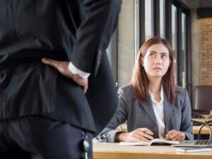 Scared employee at her office desk looking up at her angry boss berating her