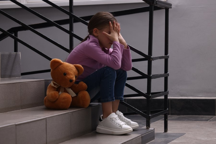 Un niño pequeño sentado en una escalera sosteniendo su cabeza en sus manos con un oso de peluche a su lado