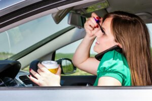 A woman is distracted by applying makeup as she drives, her second hand is holding a cup of coffee and the steering wheel at the same time