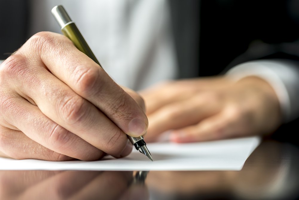 A man in a business suit holds a pen as he writes a hand-written letter on a desk.