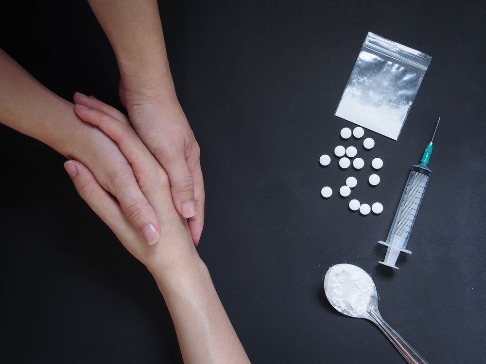 Two hands reach out to hold the hand of a drug user, on a table next to illegal drugs, pulls, a syringe, and a spoon and baggie filled with white powder