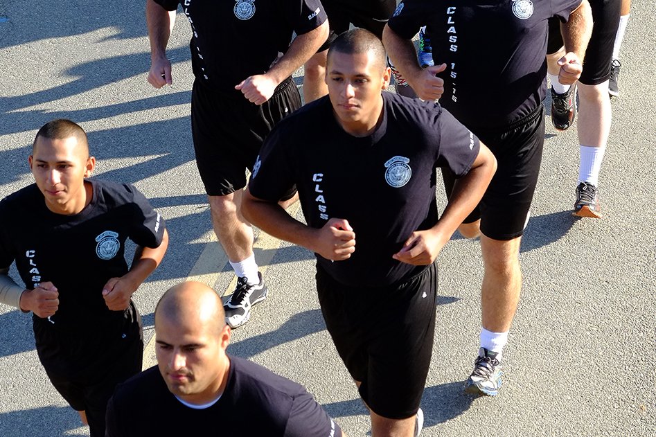 Cadetes de la academia de policía con camisetas con insignias de policía impresas, corriendo en formación en una calle de la ciudad