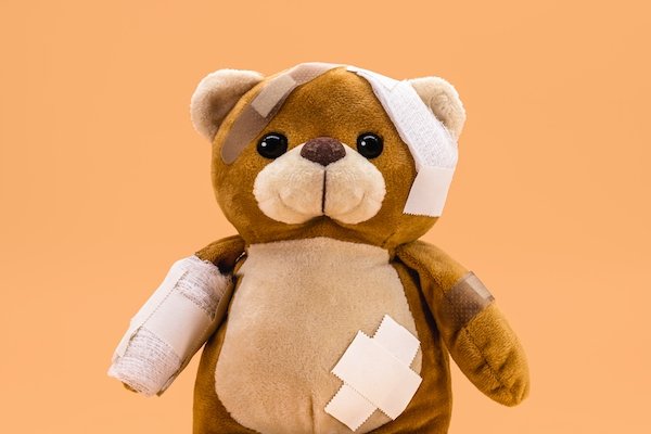 brown teddy bear with bandages on his arm, ear, and belly, against an orange background