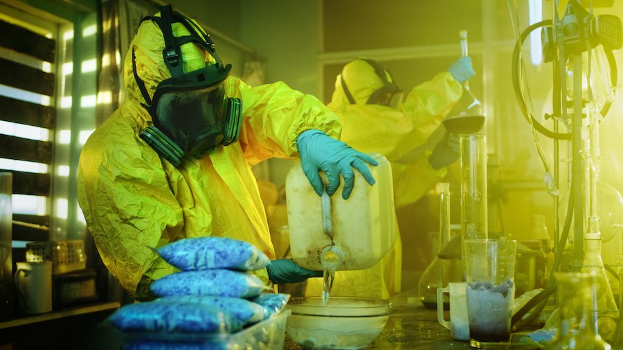 Hombre en traje de cuerpo completo "cocinando" metanfetamina en un laboratorio oscuro lleno de viales, tubos y quemadores