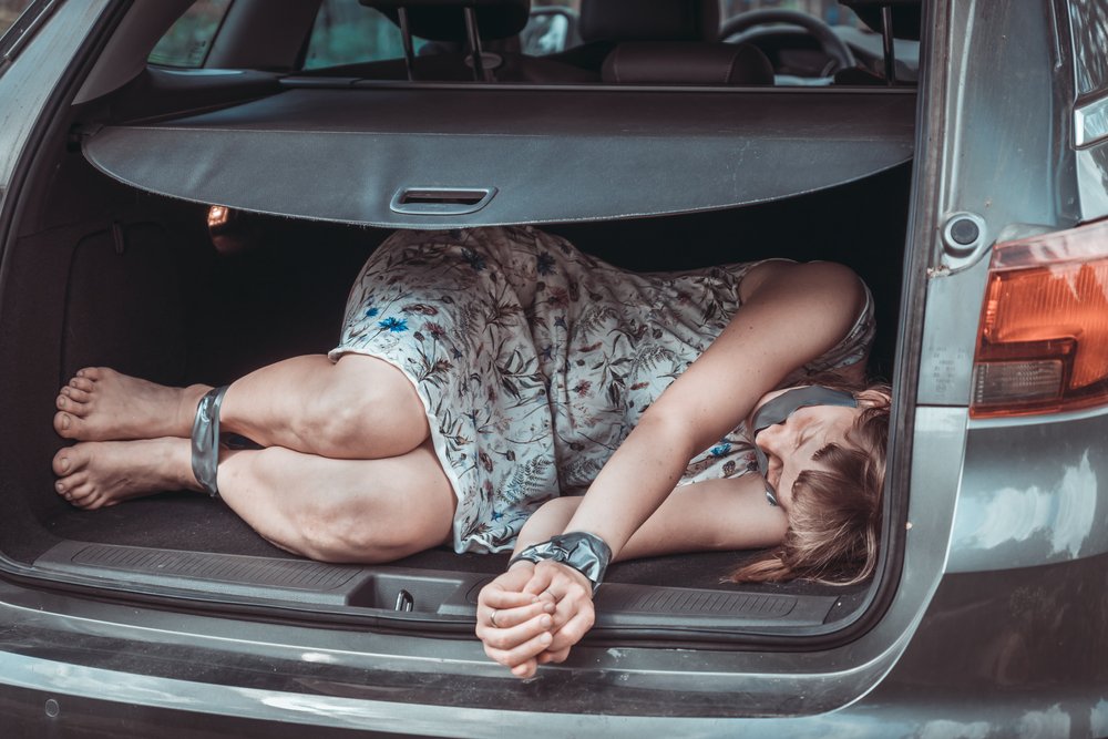 Young blonde woman who is barefoot with dirty feet, is bound and gagged with duct tape around her mouth, wrists, and ankles, tied up in the trunk of a car, she is wearing a flowery dress