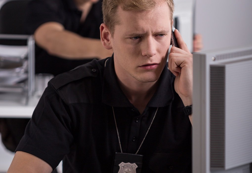 Joven investigador de policía sentado en una cabina en la estación de policía lleva una placa alrededor del cuello mientras habla por teléfono y usa una computadora