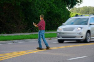 Man is using his phone as he walks across double-yellow lines in the middle of a road, a fast-moving SUV is driving towards him.