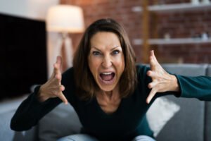 Angry, screaming woman making criminal threats at the camera and gesticulating with her hands while she sits on a couch in a living room