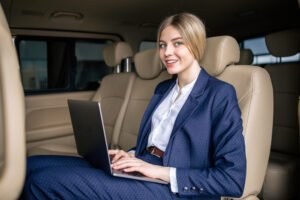 A young blonde woman wearing professional business attire is sitting in the backseat of a passenger van while she types on a laptop computer.