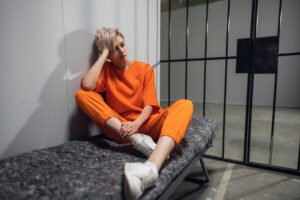 young blonde woman wearing an orange prison jumpsuit sits on a cot inside a jail cell