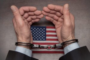 We can see the hands of a man wearing handcuffs, resting upon a table with an American flag.
