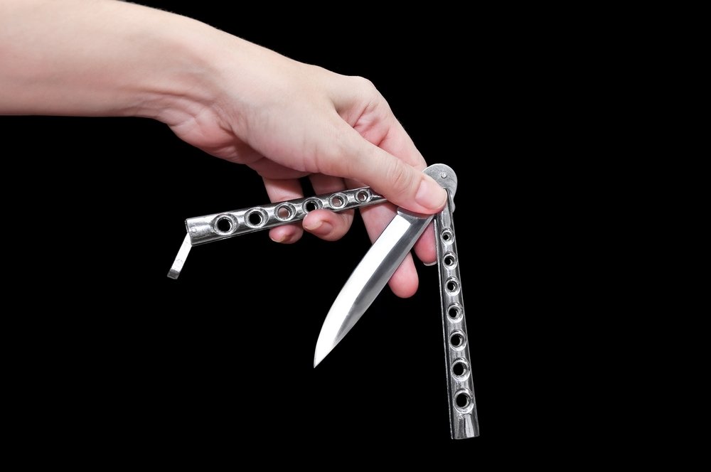 A hand holds a sharp metal blade with two metal handles that hang to either the side, one side of the handle has a clip used for closing the knife shut between the handles.
