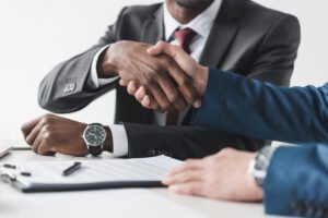 A black man in a business suit shakes the hand of a Caucasian man. They are sitting at a conference table and there is a contract and pen sitting on the table.
