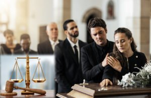 Interior: funeral home with a line of people in black suits waiting to pay their respects. A man and woman embrace hands as the woman places her other hand on the casket, in the bottom left corner of the image, the scales of justice and a gavel on the desk of a downtown law firm fade into view