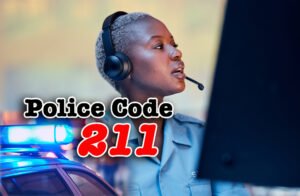 A 30 year old African-American woman in a blue police uniform talks into a communication headset as she looks at a security monitor, there is an inset in the bottom left corner of the image featuring a police car with lights and sirens and overlaid text reading "police code 211"