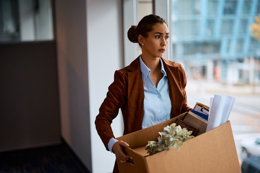 Sad-looking fired worker holding a cardboard box of office belongings as she leaves her old office