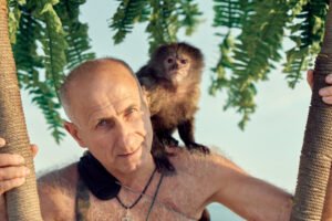 A man with a monkey on his shoulder with tree branches in the background
