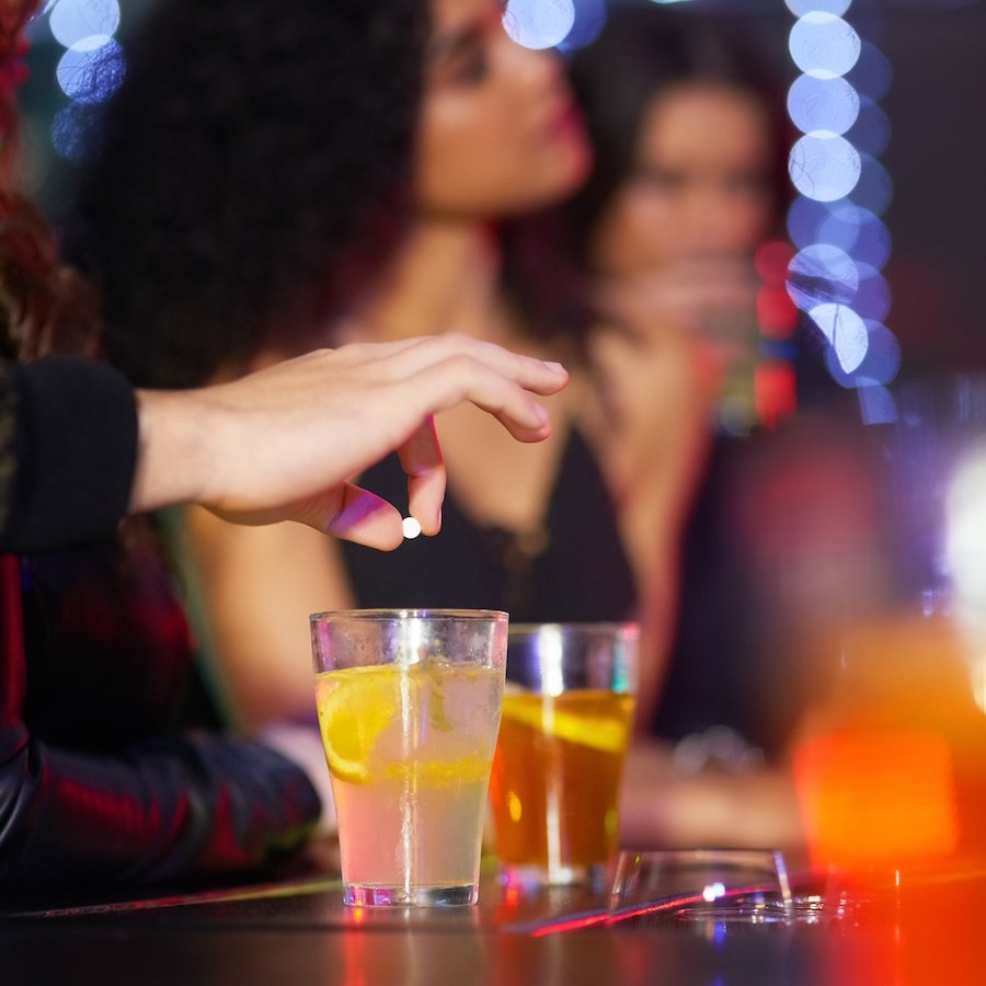Closeup de una mano a punto de dejar caer una píldora blanca en una bebida alcohólica en una mesa de bar, contra un fondo de club que muestra a dos mujeres ajenas a la droga