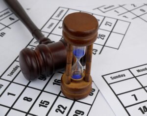A gavel and an hourglass sit atop the shuffled pages of a calendar. Judge gavel and hourglass on calendar pages background.