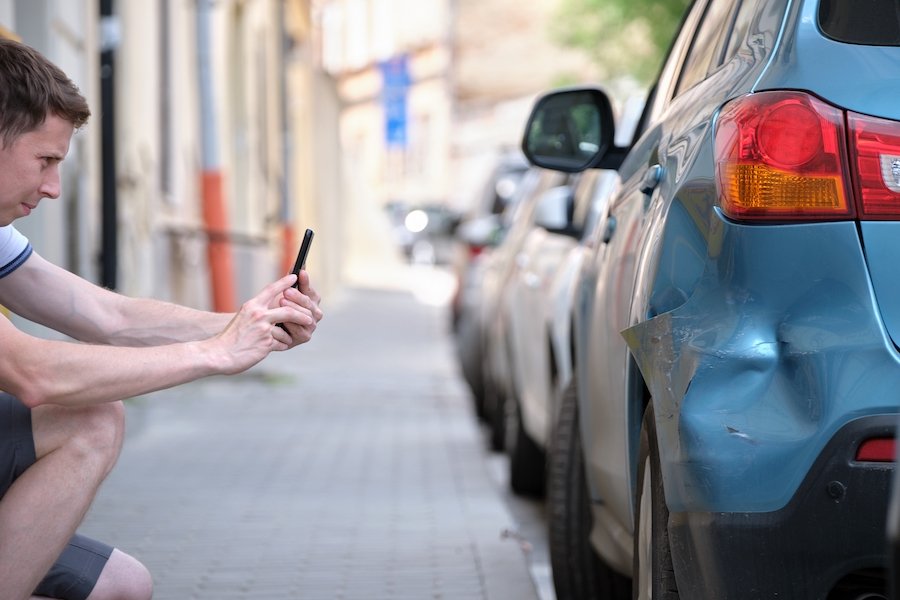 Sad looking young man taking a photo of a dented fender of a car parked on a street
