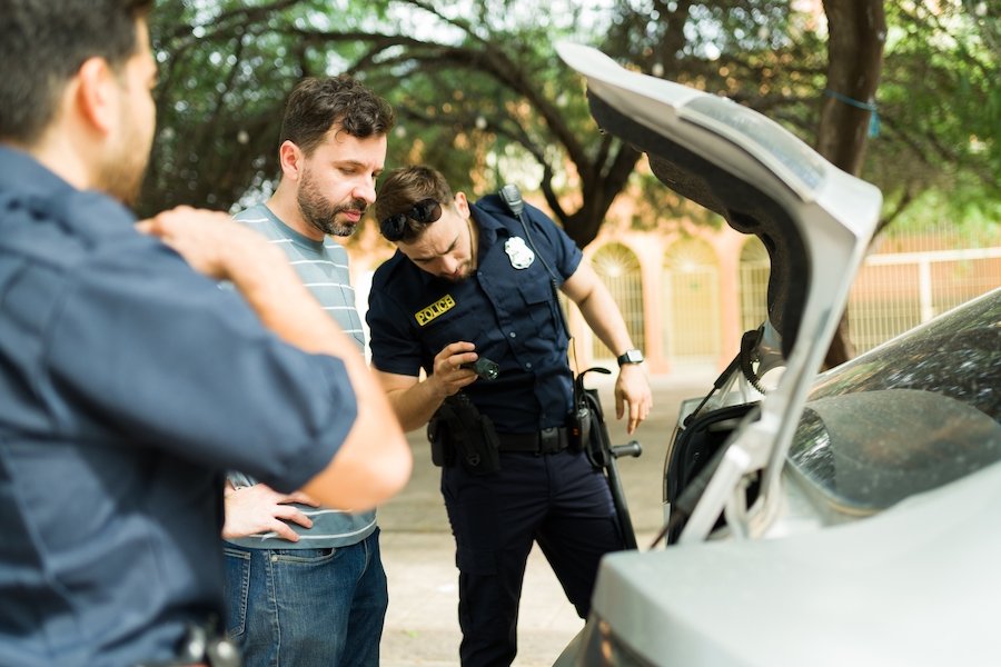 Dos oficiales de policía registrando la cajuela del automóvil mientras el conductor observa tímidamente