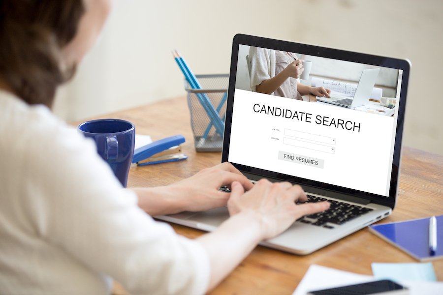 Woman at her desk typing on her laptop, and her screen says "Candidate Search"