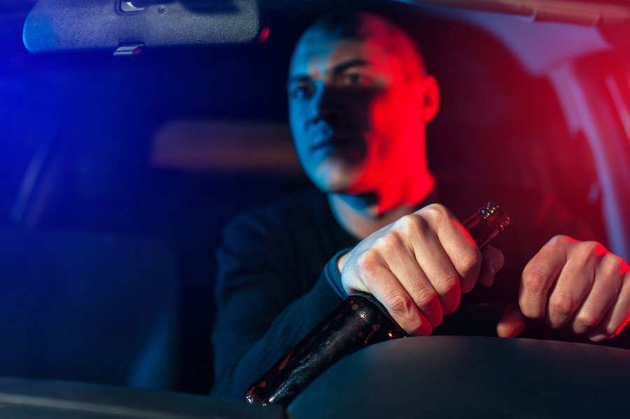 Driver holding a beer bottle while driving with police car lights visible through the rear windshield