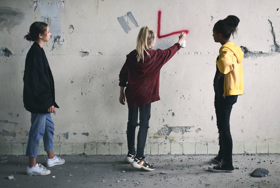 Three young teens outside during the day "tagging" a wall with graffiti