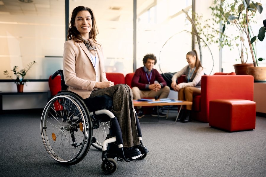 Worker in wheelchair in an office lobby, with two co-workers in the background along with office plants