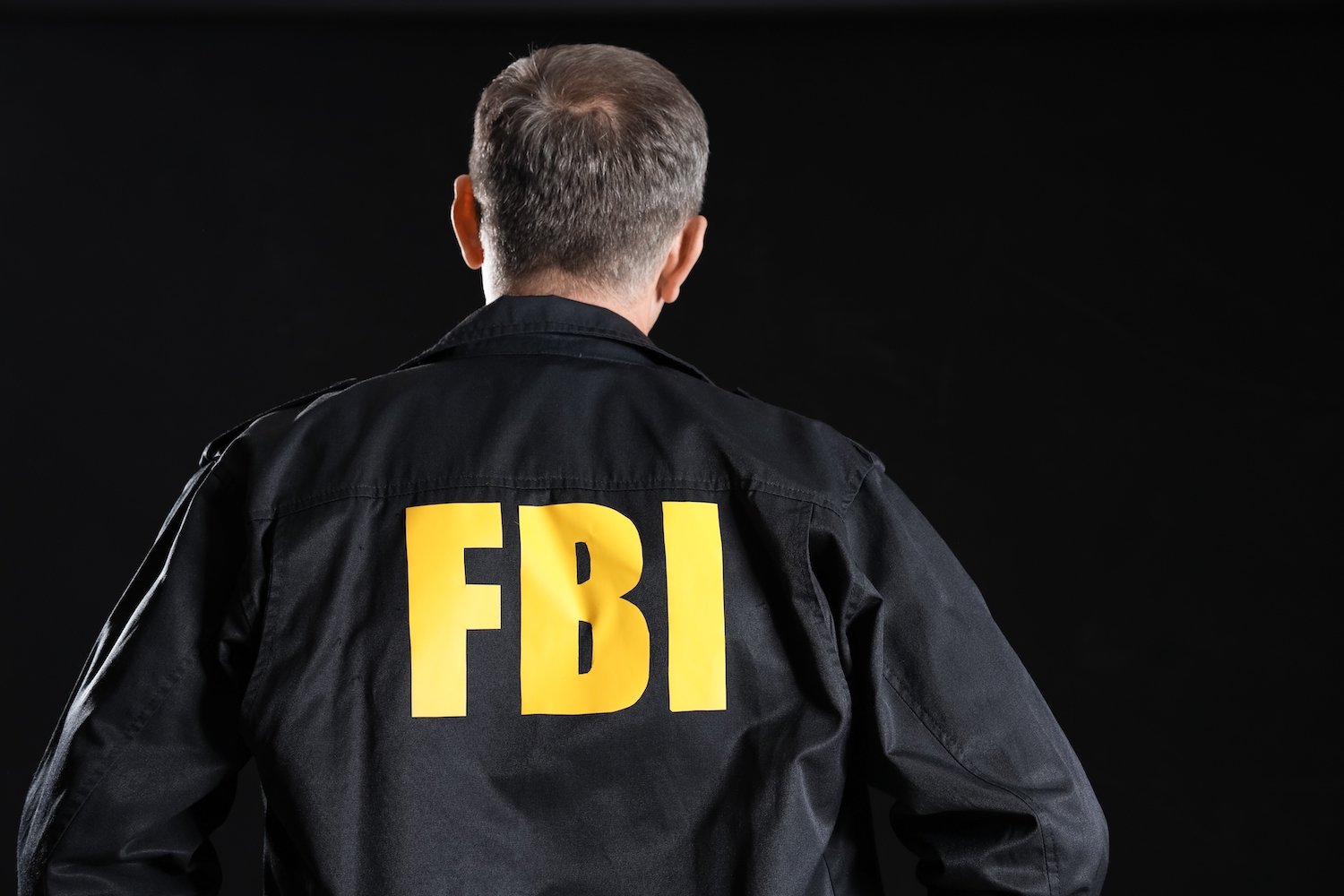 Rear view of an FBI agent wearing a black jacket with "FBI" embossed on the jacket in gold letters, against a black background