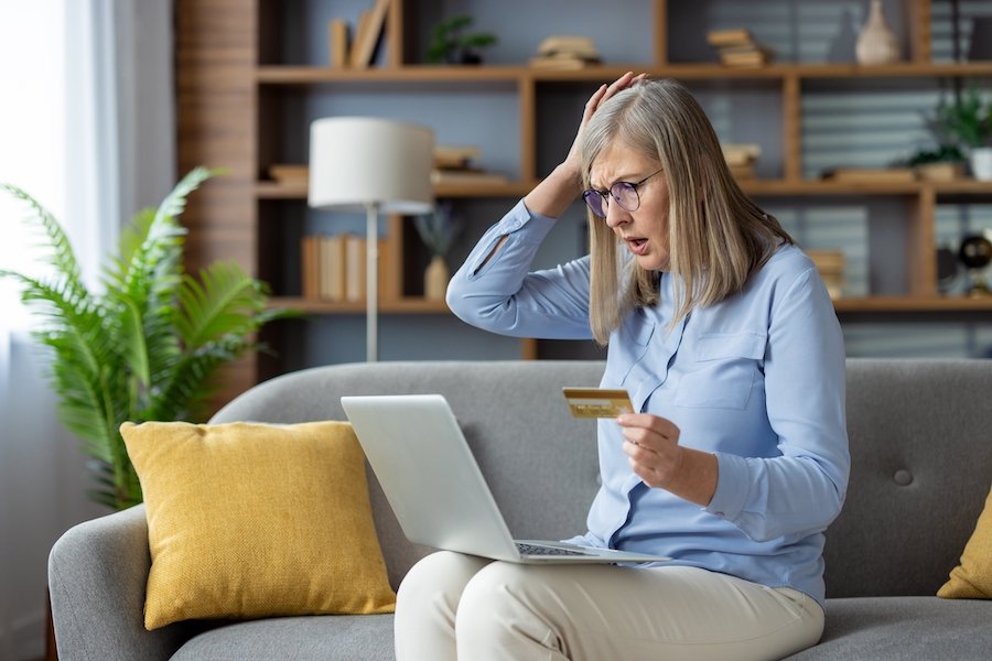 Mujer sosteniendo su cabeza en desesperación al darse cuenta de que es víctima de robo de identidad. Ella está sosteniendo su tarjeta de crédito y mirando incrédula su computadora portátil mientras está sentada en el sofá.