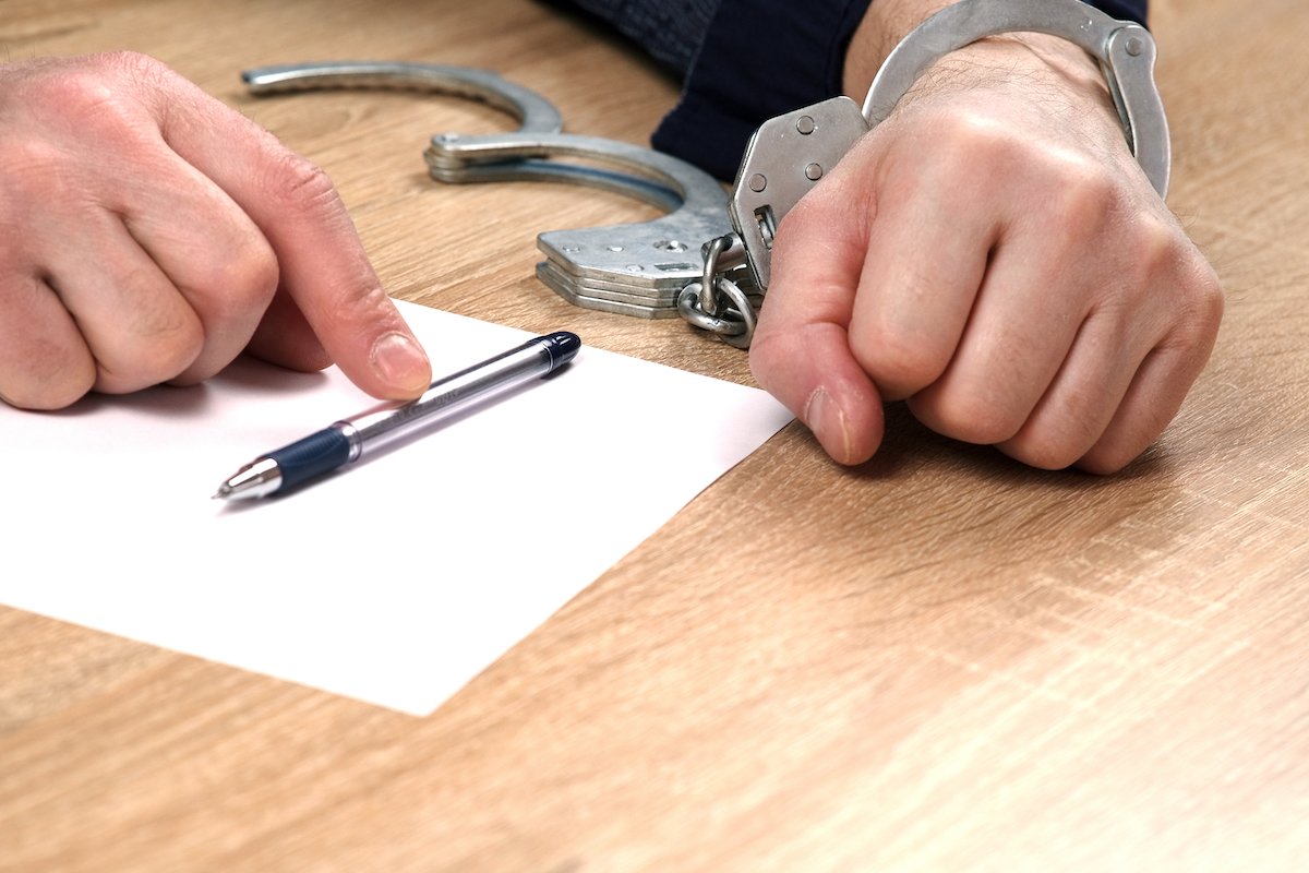 Primer plano de un hombre con esposas en una sola muñeca, firmando un documento con la otra mano prometiendo presentarse a futuras audiencias judiciales