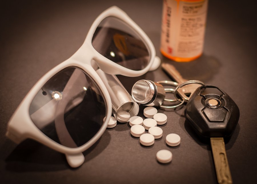 Car key next to a prescription pill bottle and pills and a pair of sunglasses