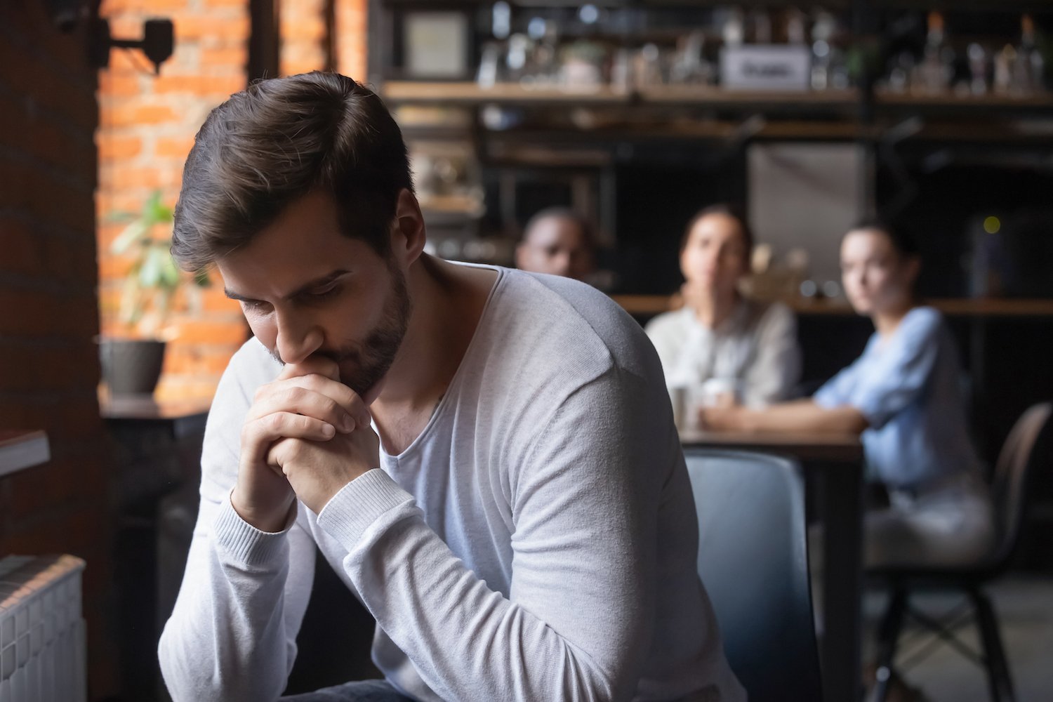 Hombre en cafetería luciendo abatido mientras personas en una mesa cercana lo miran con juicio