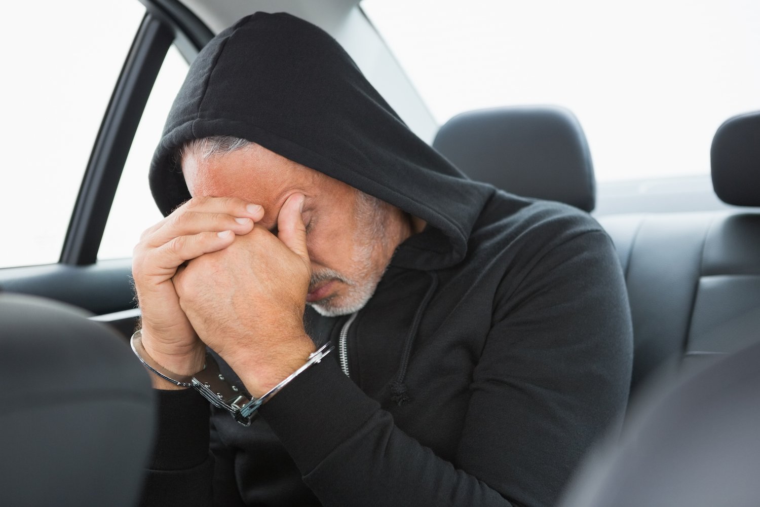 Man in black hoodie holding his head in his hands, which are handcuffed, as he sits in a police vehicle