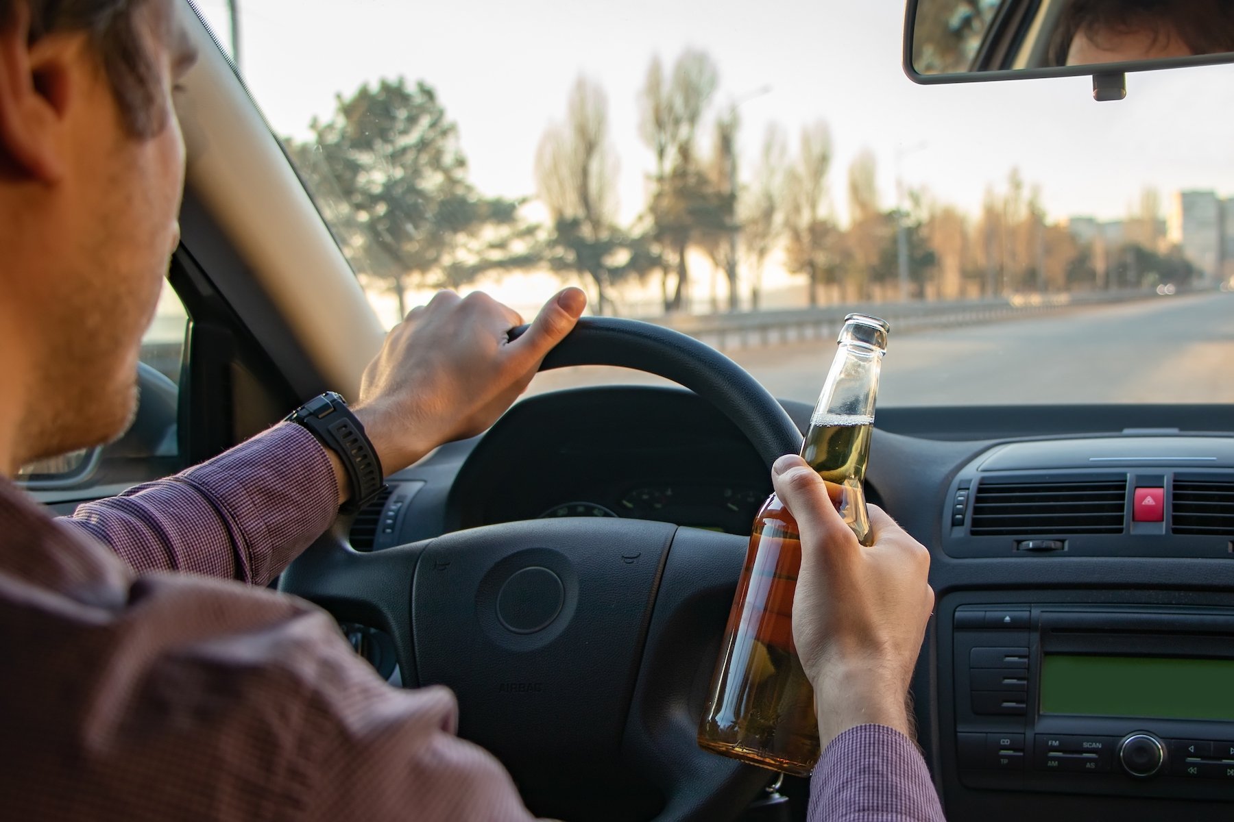 Driver holding steering wheel in one hand and a bottle of beer in the other