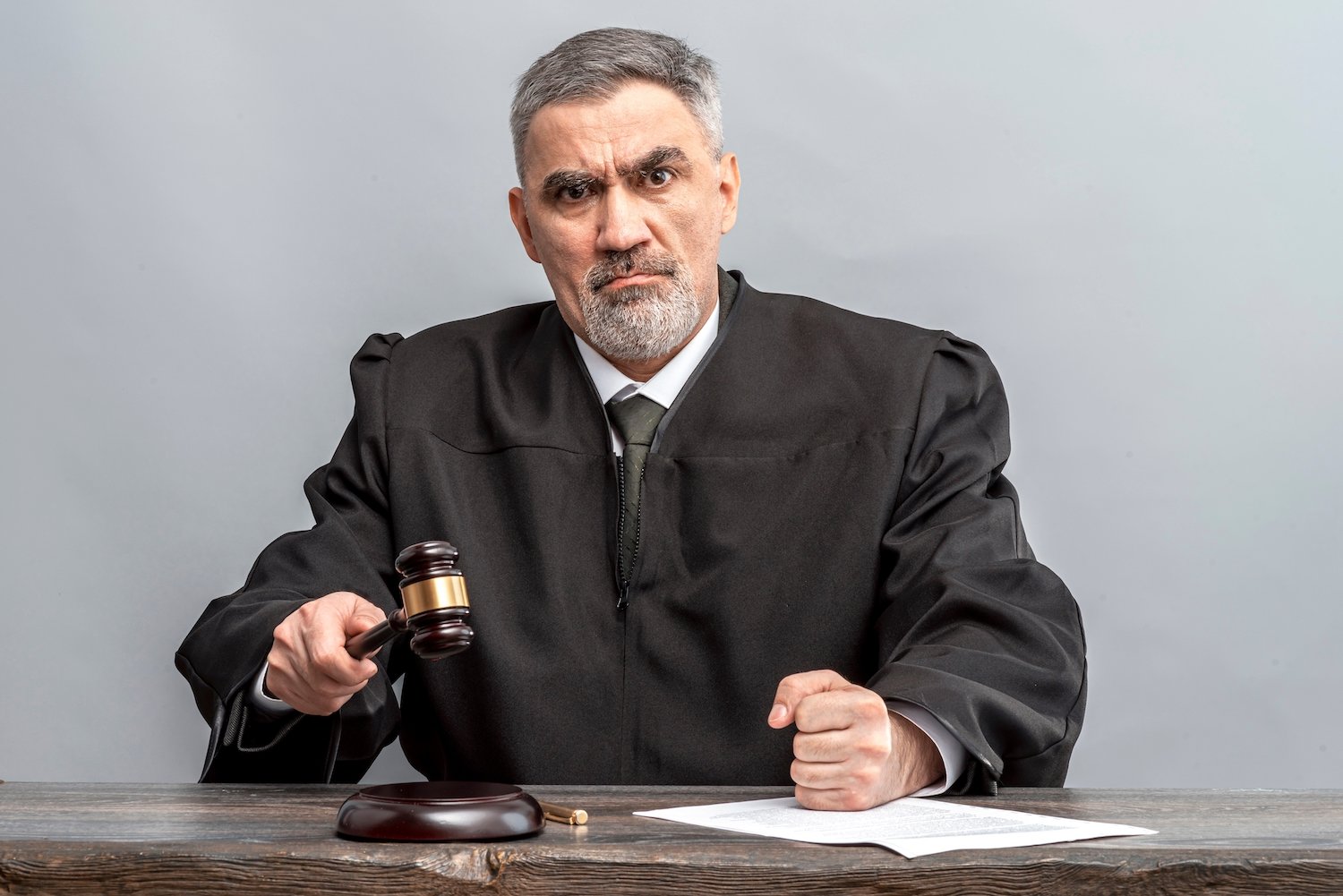 Judge in a black robe at a desk hammering down a gavel against a grey wall