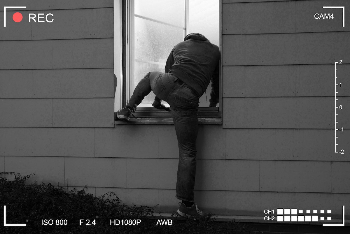 Rear view of a burglar entering a house through an open window as an example of 2nd-degree burglary