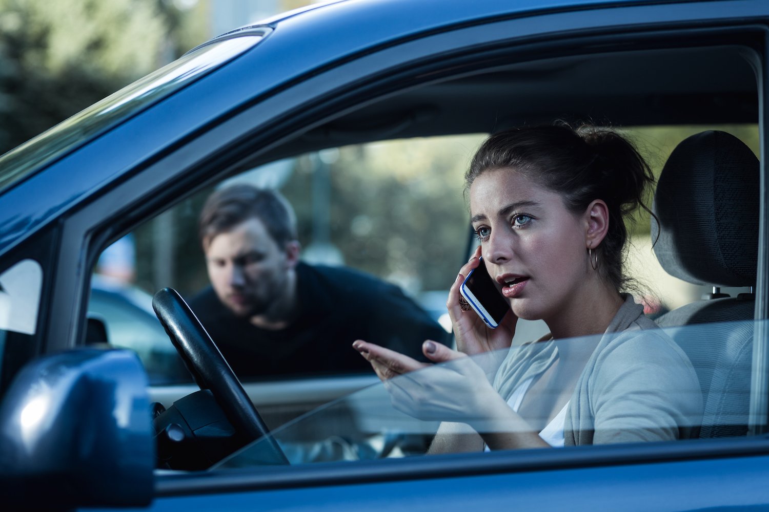 Mujer hablando por teléfono en su auto mientras un hombre la acosa a través de una ventana