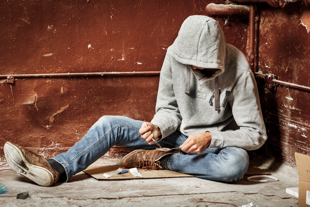 Hooded addict on a street corner administering drugs to himself