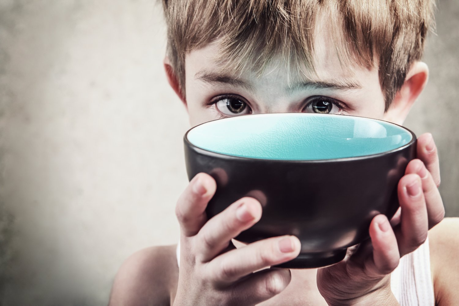 Hungry, neglected young boy holding up an empty bowl against a grey background