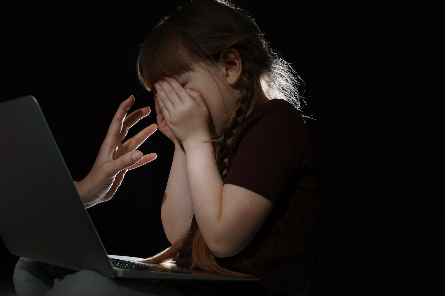 Stranger reaching frightened little child through a laptop screen against a dark background. 