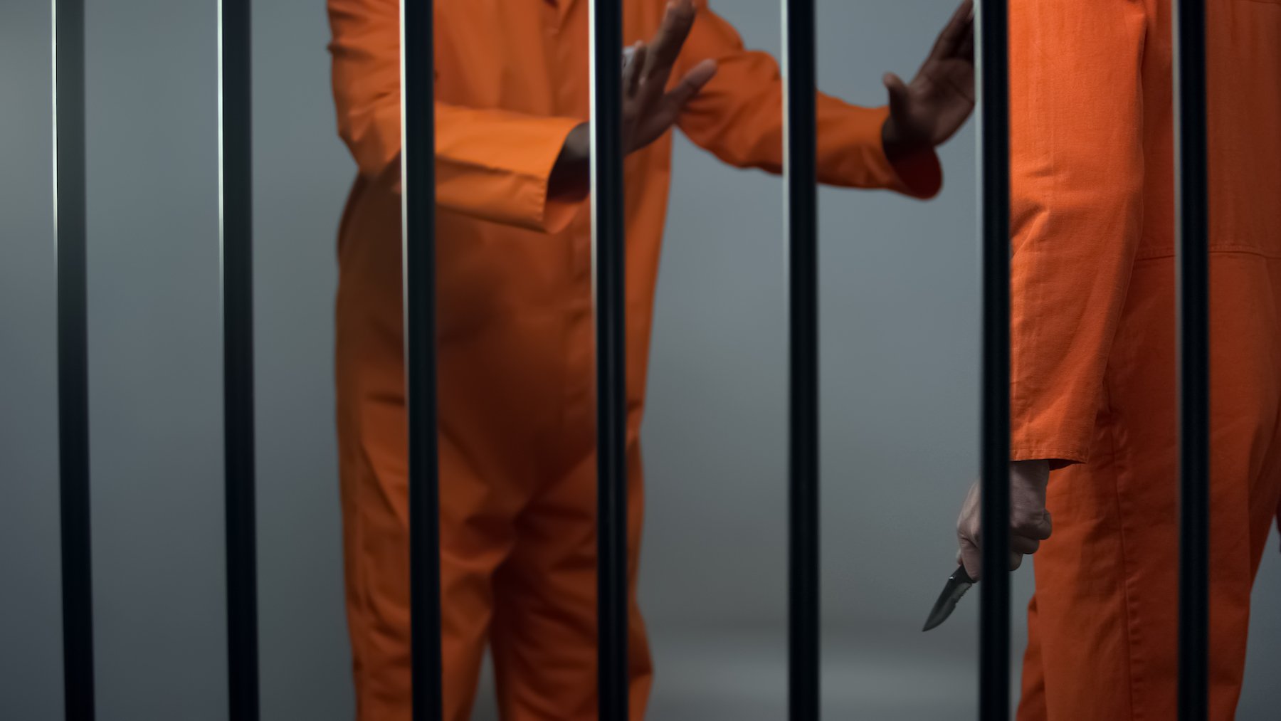 Dos reclusos con uniforme naranja detrás de rejas, y uno sostiene un cuchillo